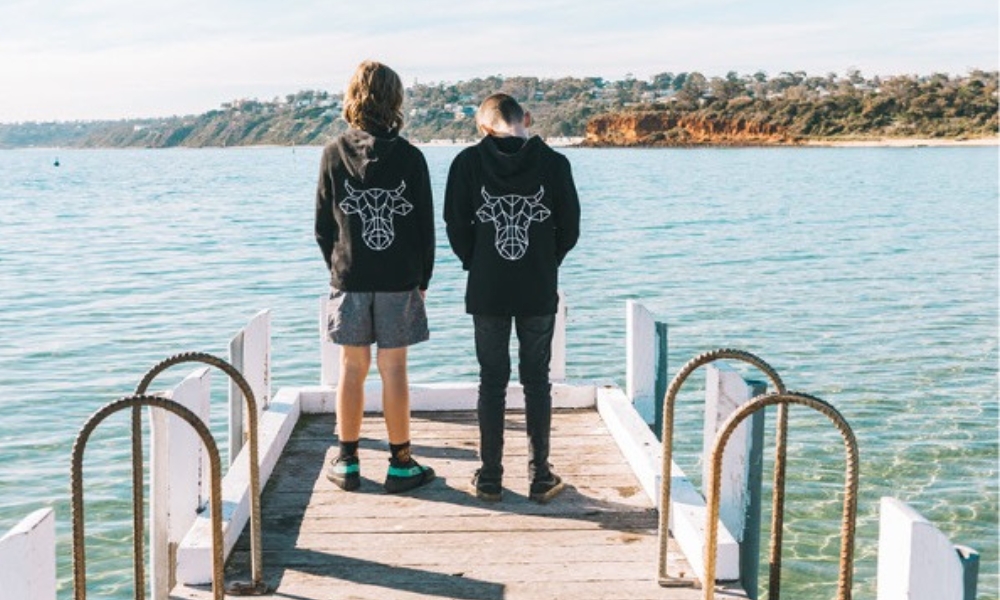 Two young boys faced away from the camera wearing jumpers with The Winey Cow logo. They are on a wooden pier with blue water surrounding them.