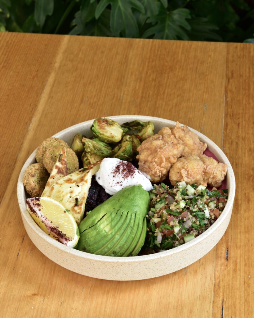 Healthy protein bowl featuring crispy chicken, falafel, grilled halloumi, smashed avocado, Brussels sprouts, tabbouleh, a dollop of labneh, lemon wedge, and roasted beetroot, served in a ceramic bowl on a wooden table.