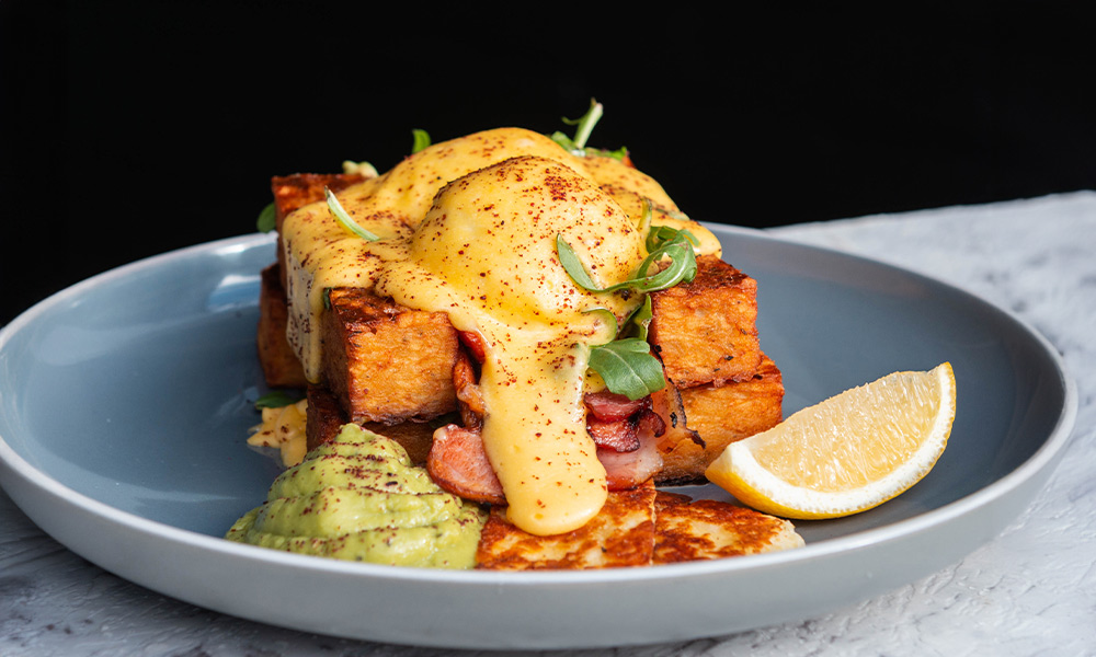 A blue plate on a marble table of a breakfast egg stack with hollandaise sauce dripping off the side.