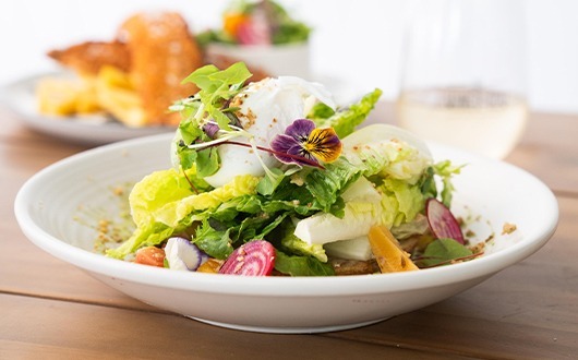 A colorful salad featuring gem lettuce, pickled beets, a poached egg and topped with edible flowers that add pops of color in shades of purple and yellow, creating an artistic and appetizing presentation.