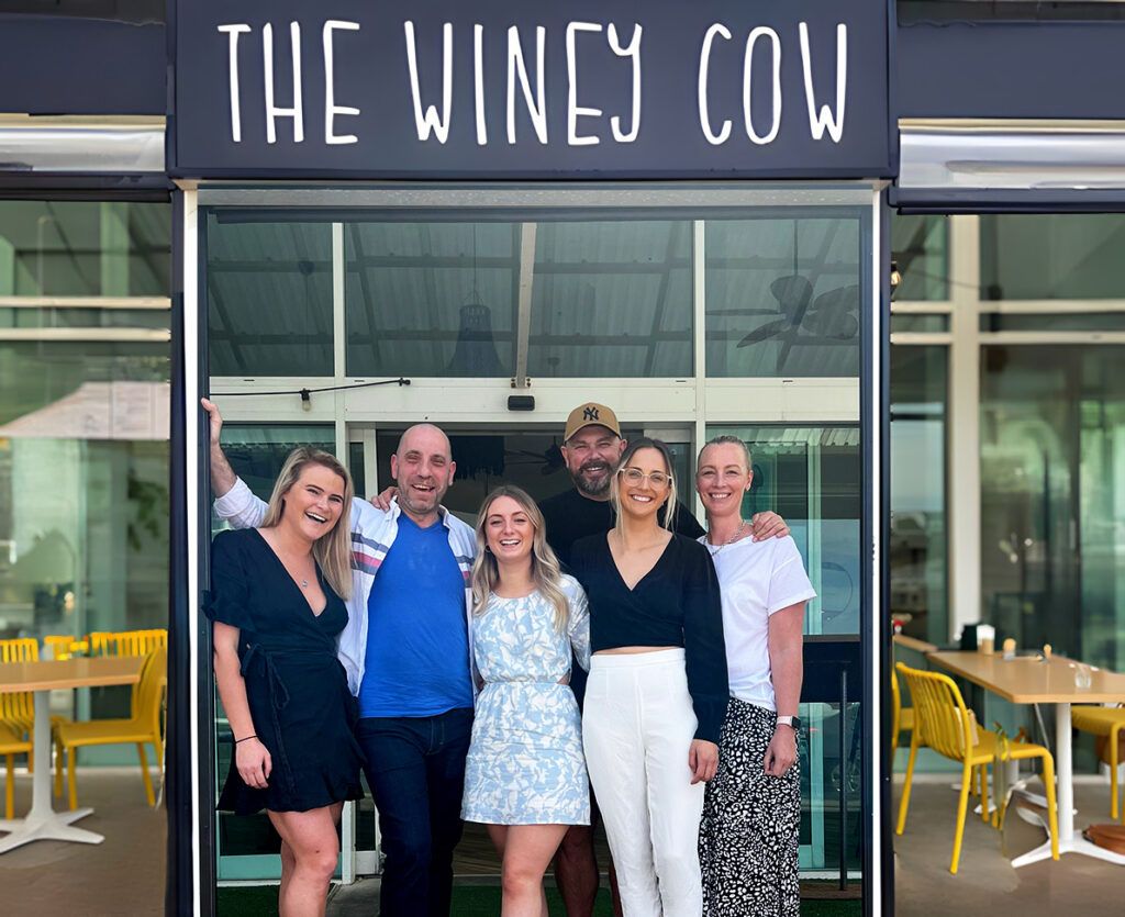 Group photo of the six owners standing in front of The Winey Cow entrance, smiling and posing together. The restaurant's sign is visible above them, and the interior with yellow chairs and tables can be seen through the open doors.