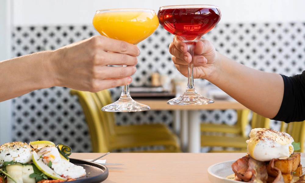 Two people clink cocktail glasses—one with an orange mimosa and the other with a red spritz—over a brunch table featuring gourmet dishes with poached eggs, bacon, and fresh vegetables at The Winey Cow.