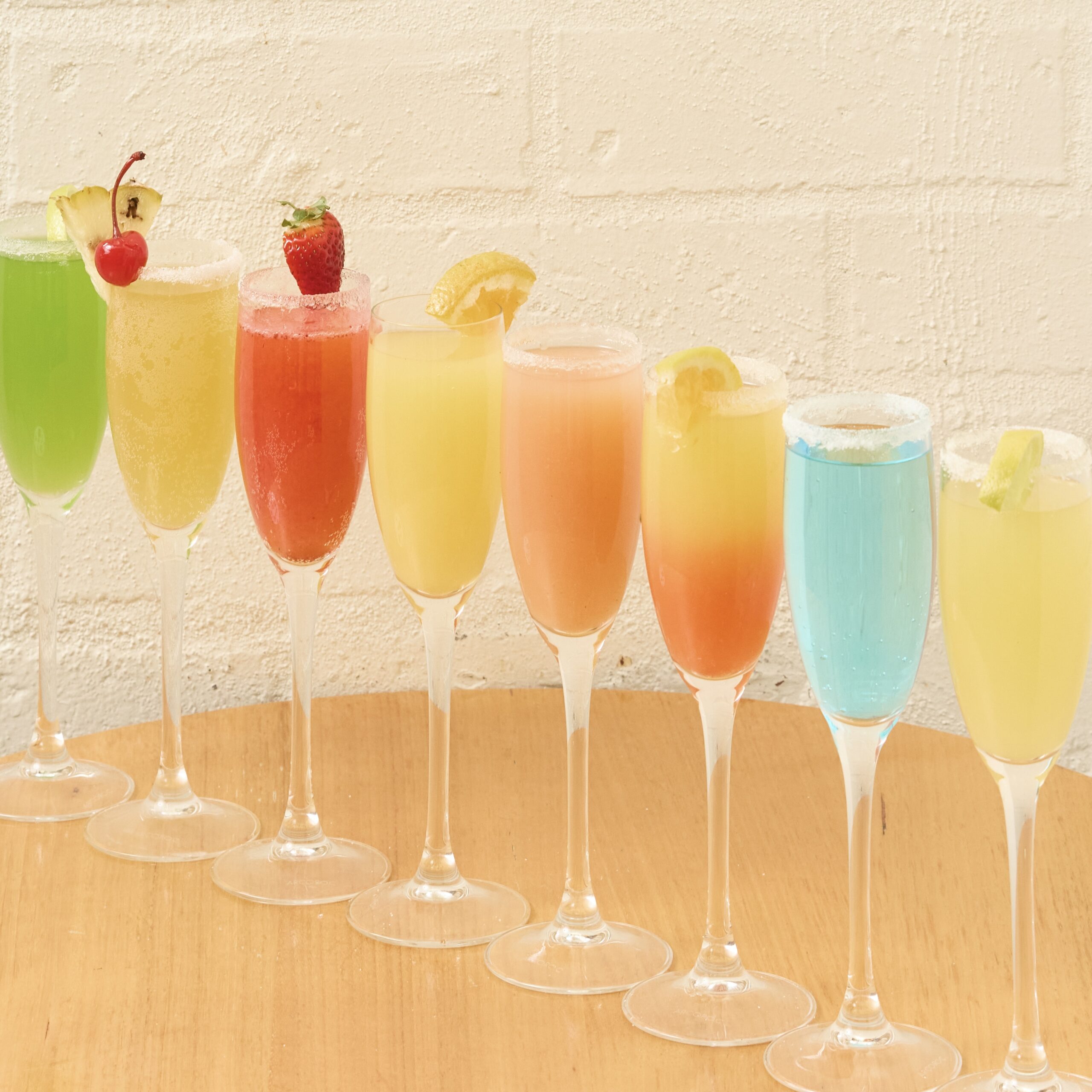 A colourful lineup of eight mimosas from The Winey Cow’s Premium Mimosa Bar, featuring vibrant flavours like Piña Colada, Strawberry Bliss, Tequila Sunrise, and more, served in champagne flutes on a wooden table against a white brick wall.