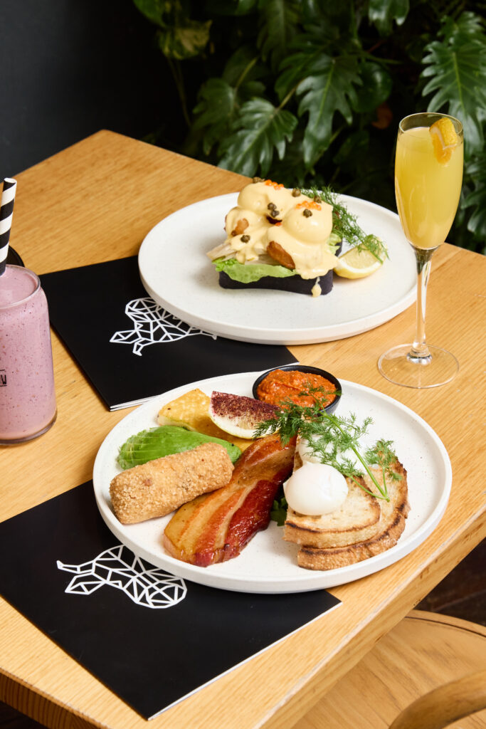 A vibrant brunch spread at The Winey Cow featuring a gourmet big breakfast with bacon, avocado, poached eggs, toast, and croquette, paired with a berry smoothie and a fresh eggs benedict dish served with a mimosa.