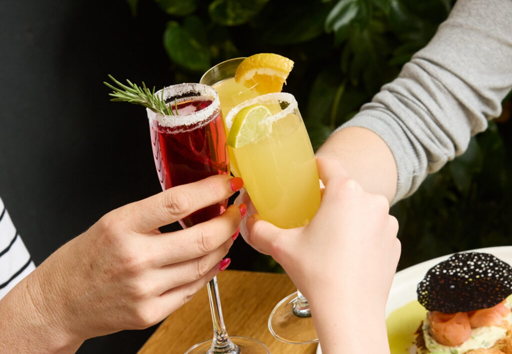 Guests toasting with vibrant mimosas during Boozy Brunch at The Winey Cow, surrounded by colourful brunch dishes and a lively, relaxed atmosphere.