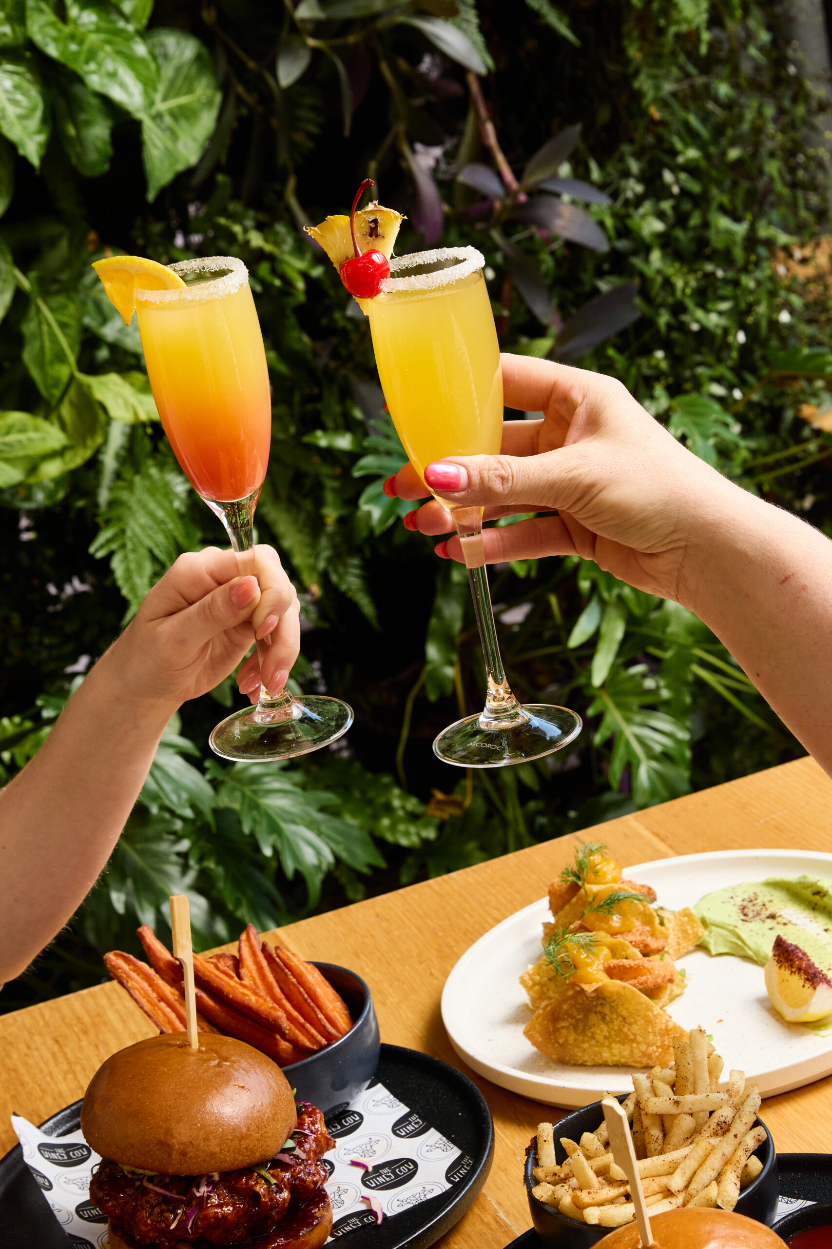 Two guests clinking tropical mimosas during Boozy Brunch at The Winey Cow, with a spread of gourmet brunch dishes including a crispy chicken burger with sweet potato fries, wonton tacos, and seasoned fries, set against a lush green backdrop.