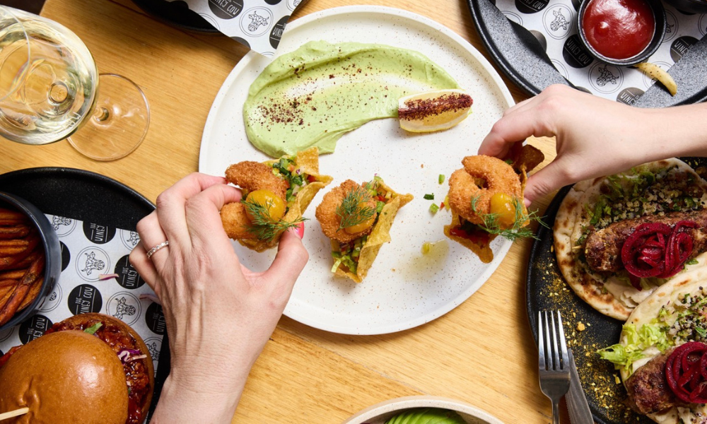 Two hands reaching for crispy fried shrimp tacos served on a white plate with avocado mousse and lemon, surrounded by a vibrant brunch spread including burgers, sweet potato fries, sausages on flatbread, and a glass of white wine