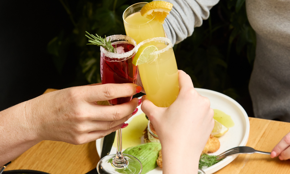 Three people clink cocktail glasses—one with an orange mimosa and the other with a red spritz—over a brunch table featuring gourmet dishes with poached eggs, bacon, and fresh vegetables at The Winey Cow.