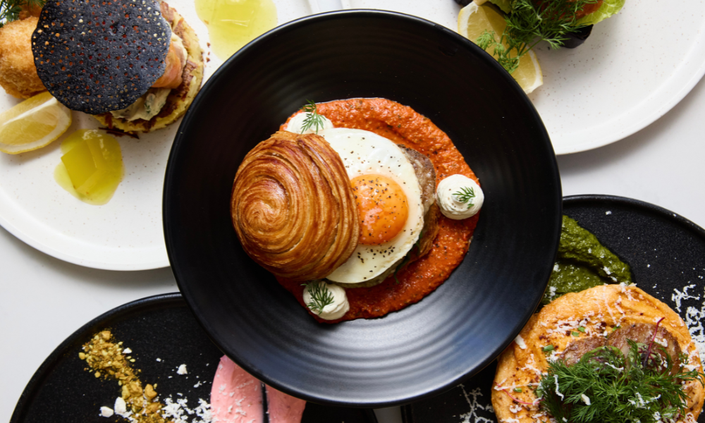 A gourmet brunch spread featuring a croissant-topped dish with a sunny-side-up egg on a bed of red pepper sauce, surrounded by various colourful and artfully plated dishes including greens, lemon wedges, and textured garnishes.