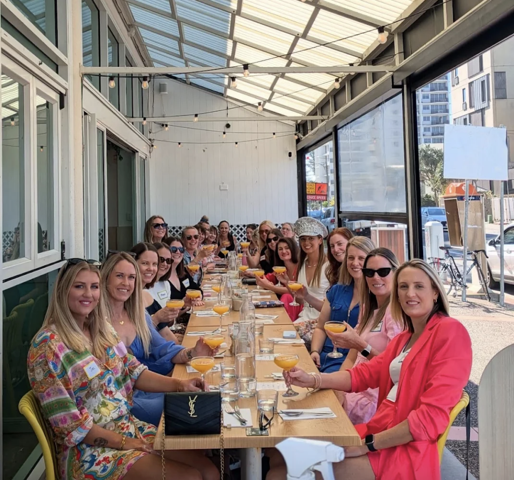 A large group of women enjoying Boozy Brunch at The Winey Cow Main Beach, seated along a long outdoor table with mimosas in hand under a covered patio, celebrating in a vibrant, beachside setting.