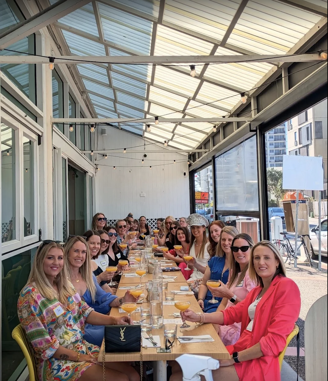 A large group of women enjoying Boozy Brunch at The Winey Cow Main Beach, seated along a long outdoor table with mimosas in hand under a covered patio, celebrating in a vibrant, beachside setting.